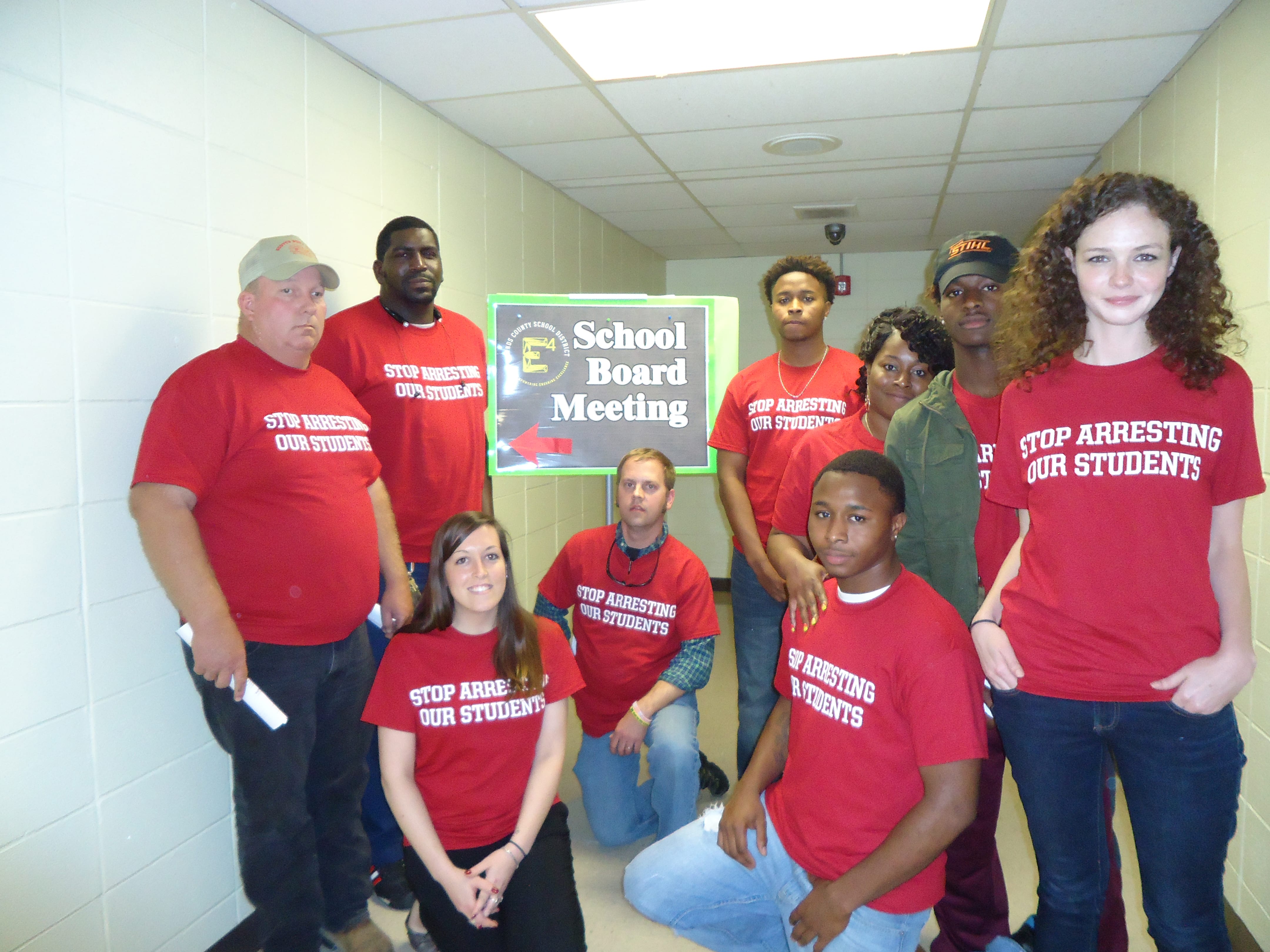 Group of people standing/sitting with red shirts reading "Stop Arresting Our Students"