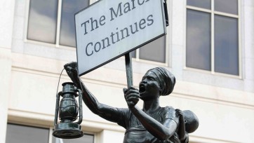 blank slate monument statue holds sign that says 'The March Continues'