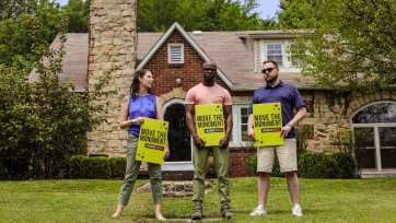 Three people holding signs that read "Move the Monument" on front lawn of home.