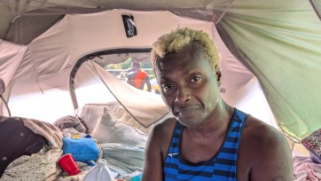A person sits inside a tent filled with sleeping gear and personal property.