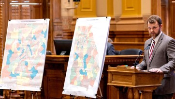Person behind podium with two maps divided with county outlines on easels