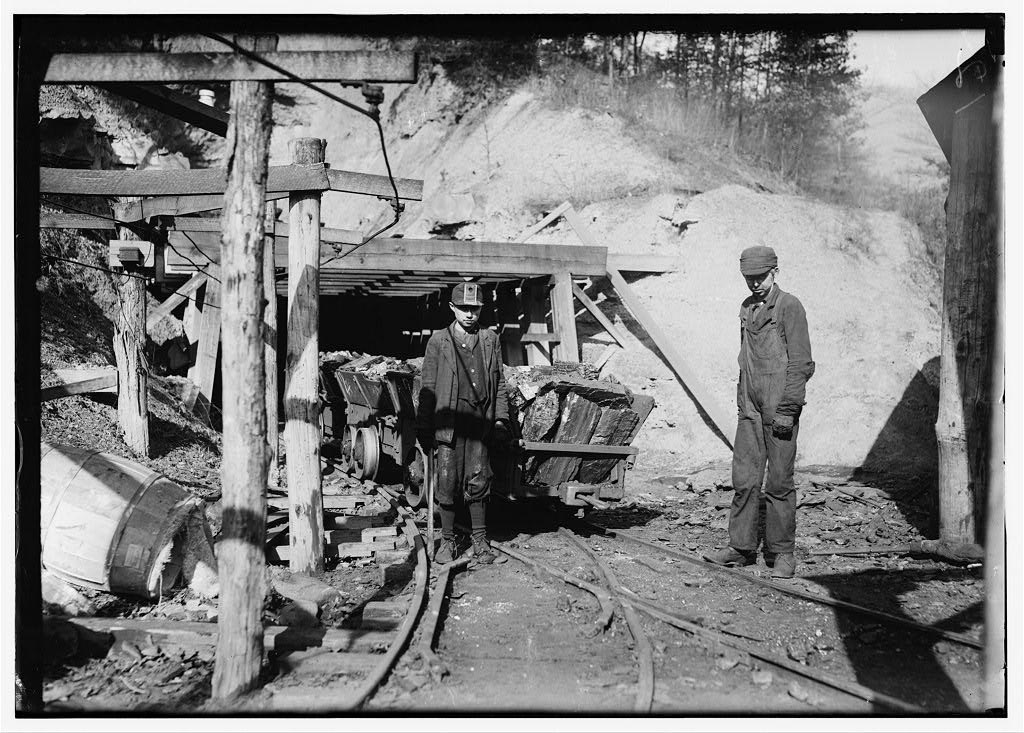 Miners stand in front of shaft opening blocked by rail car.