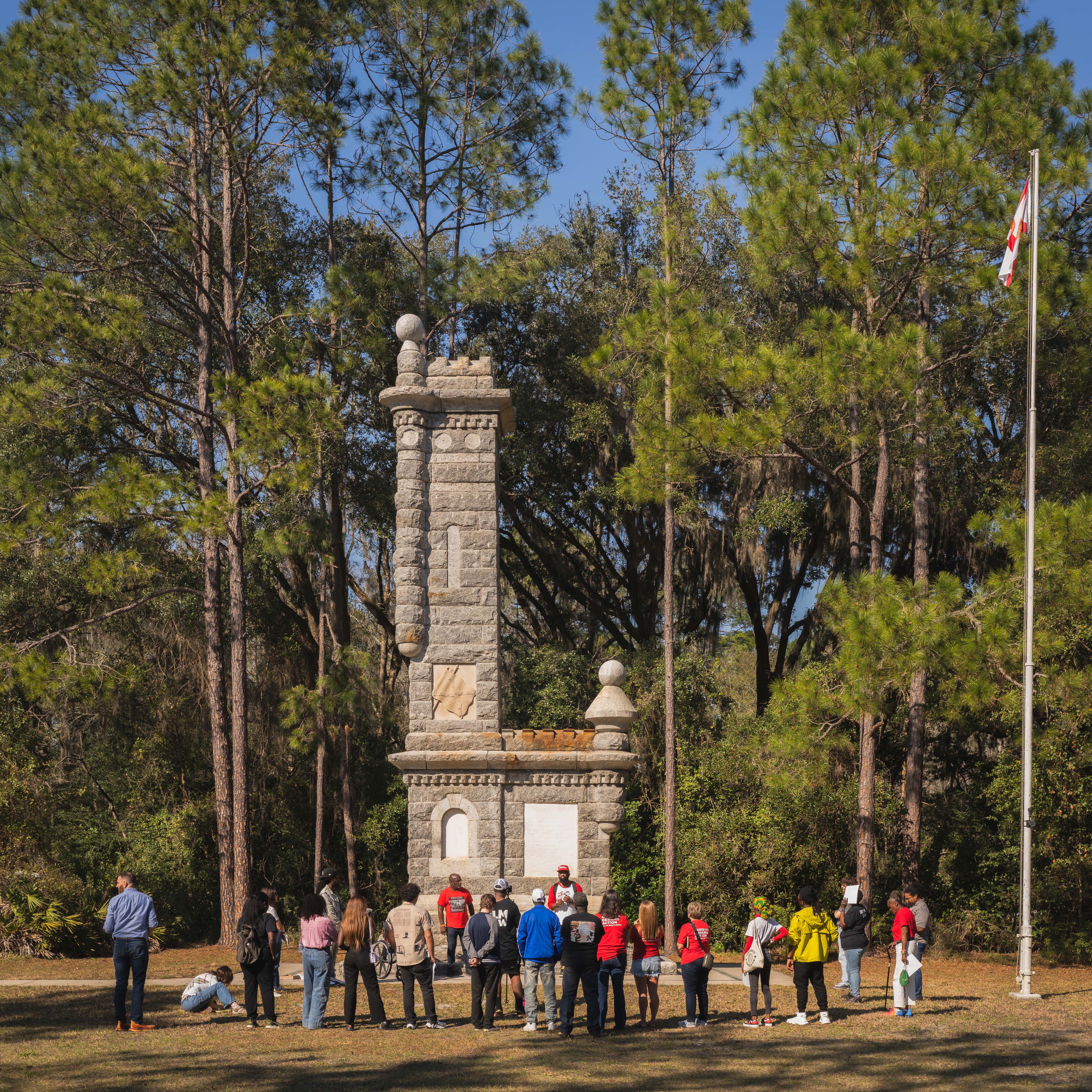 Group of people stand in front of a monument in a park setting with trees in background.