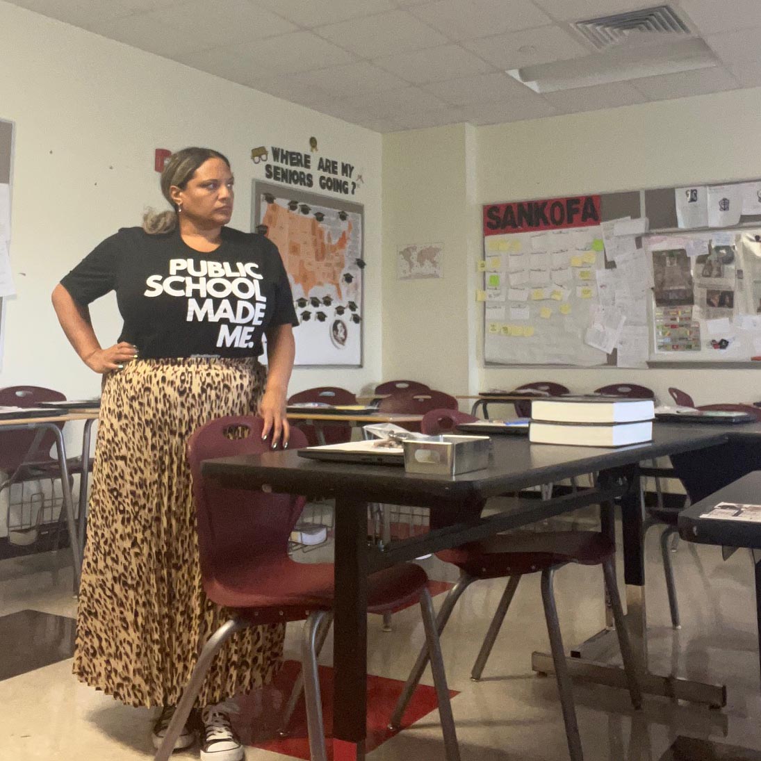 Person in a black shirt that reads "public schools made me" stands in an empty classroom.