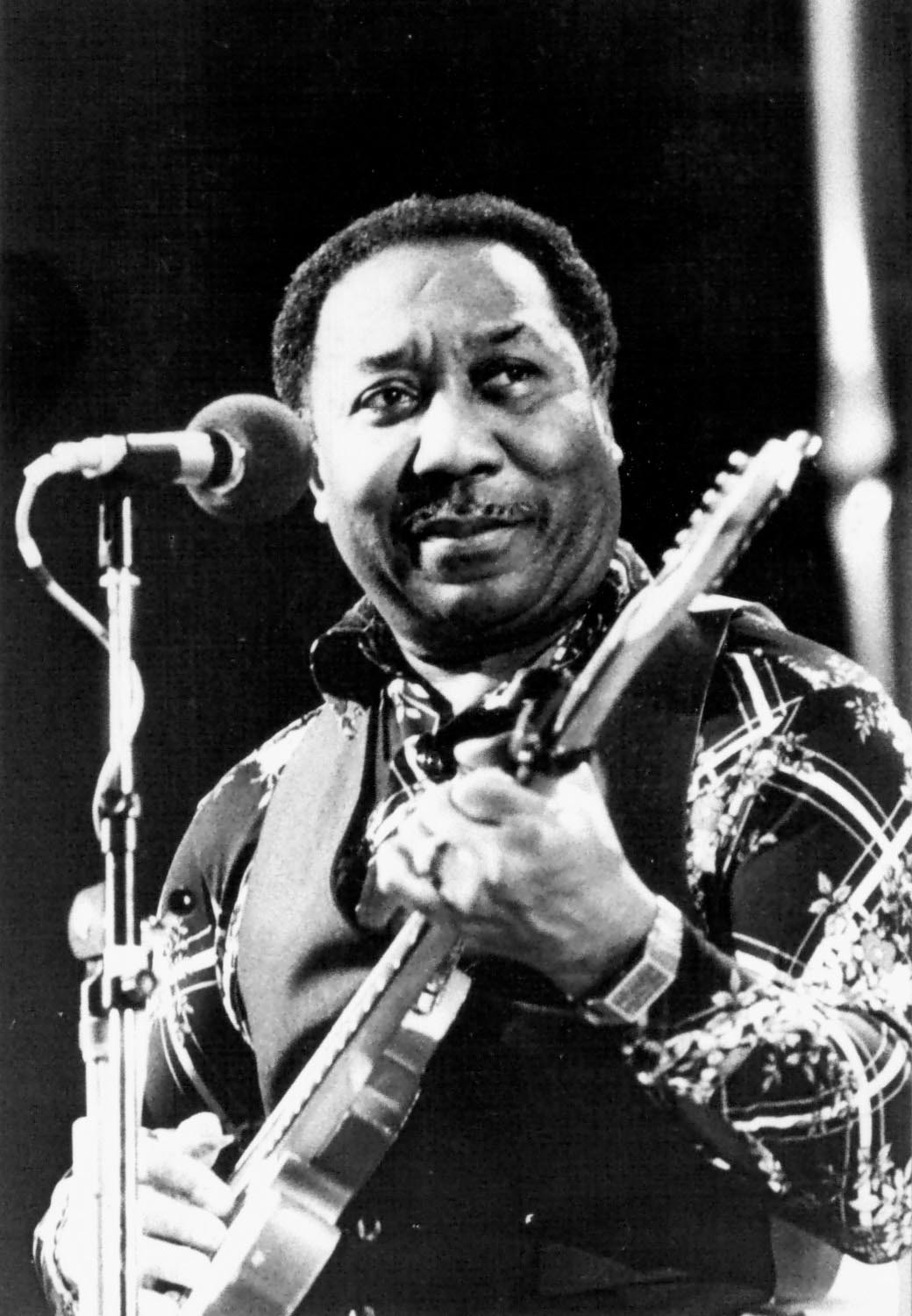 Muddy Waters at microphone strums an electric guitar.