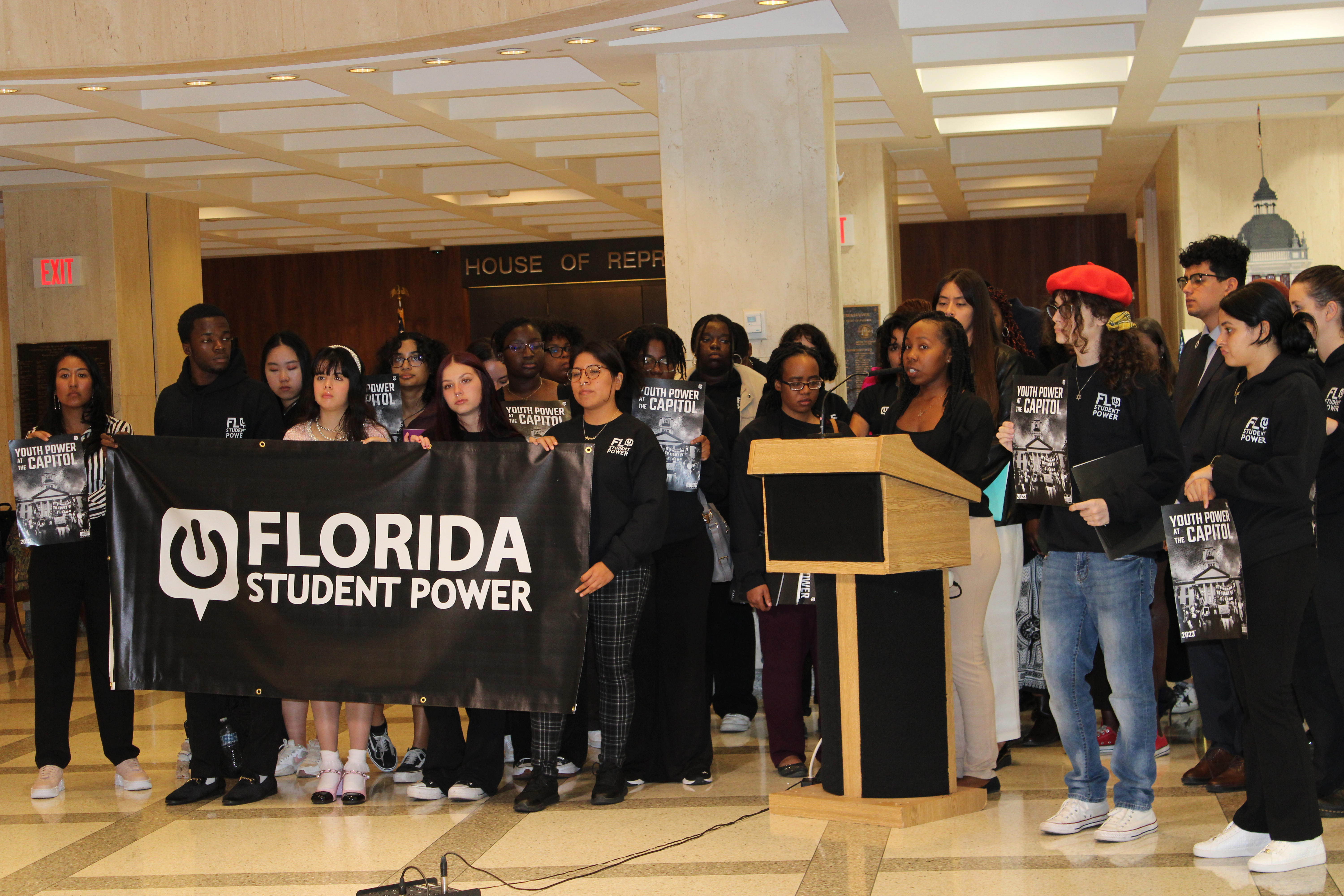 Group holds banner and signs for Florida Student Power as speaker at podium addresses open room