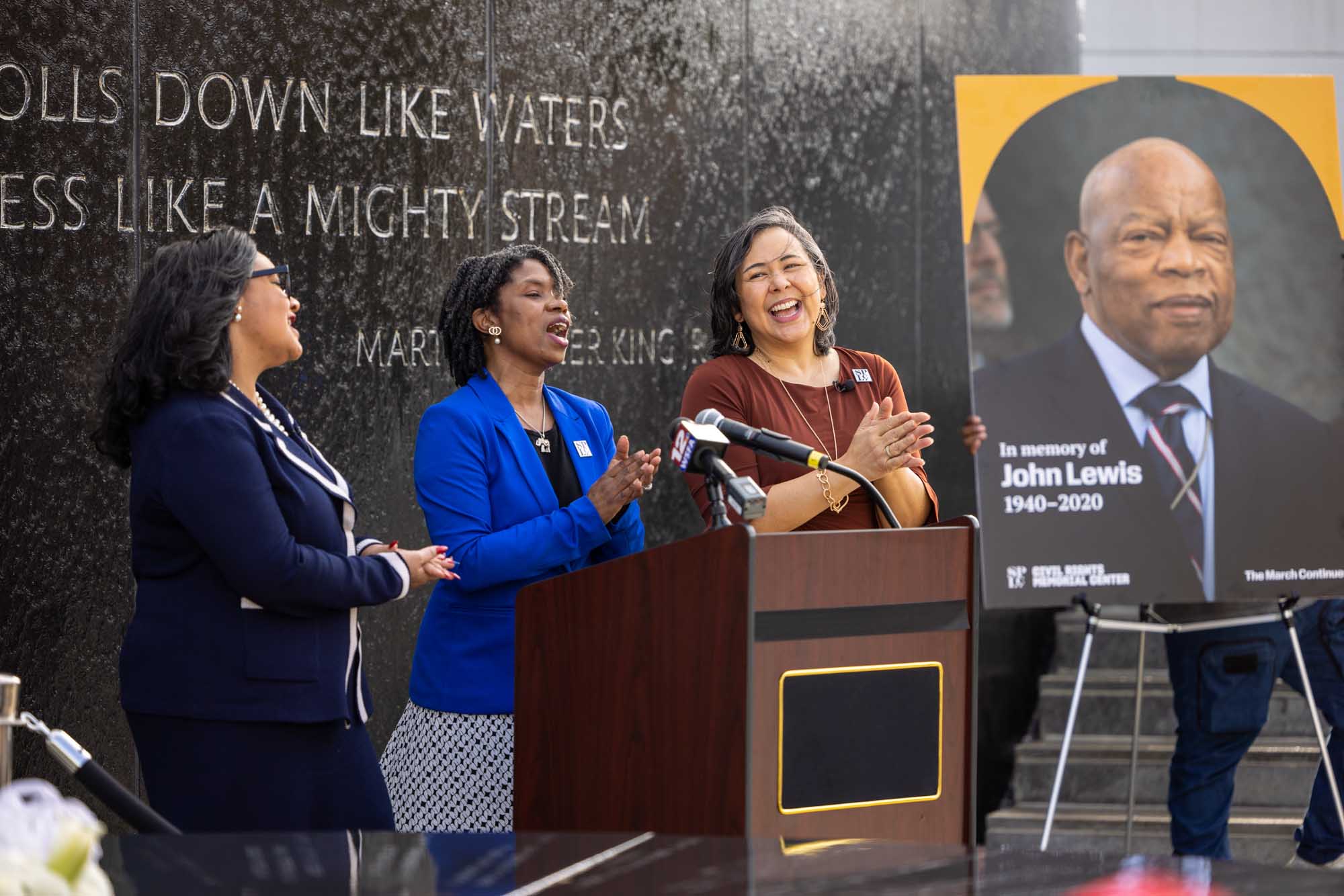 Three people lead crowd in singing before portrait of John Lewis