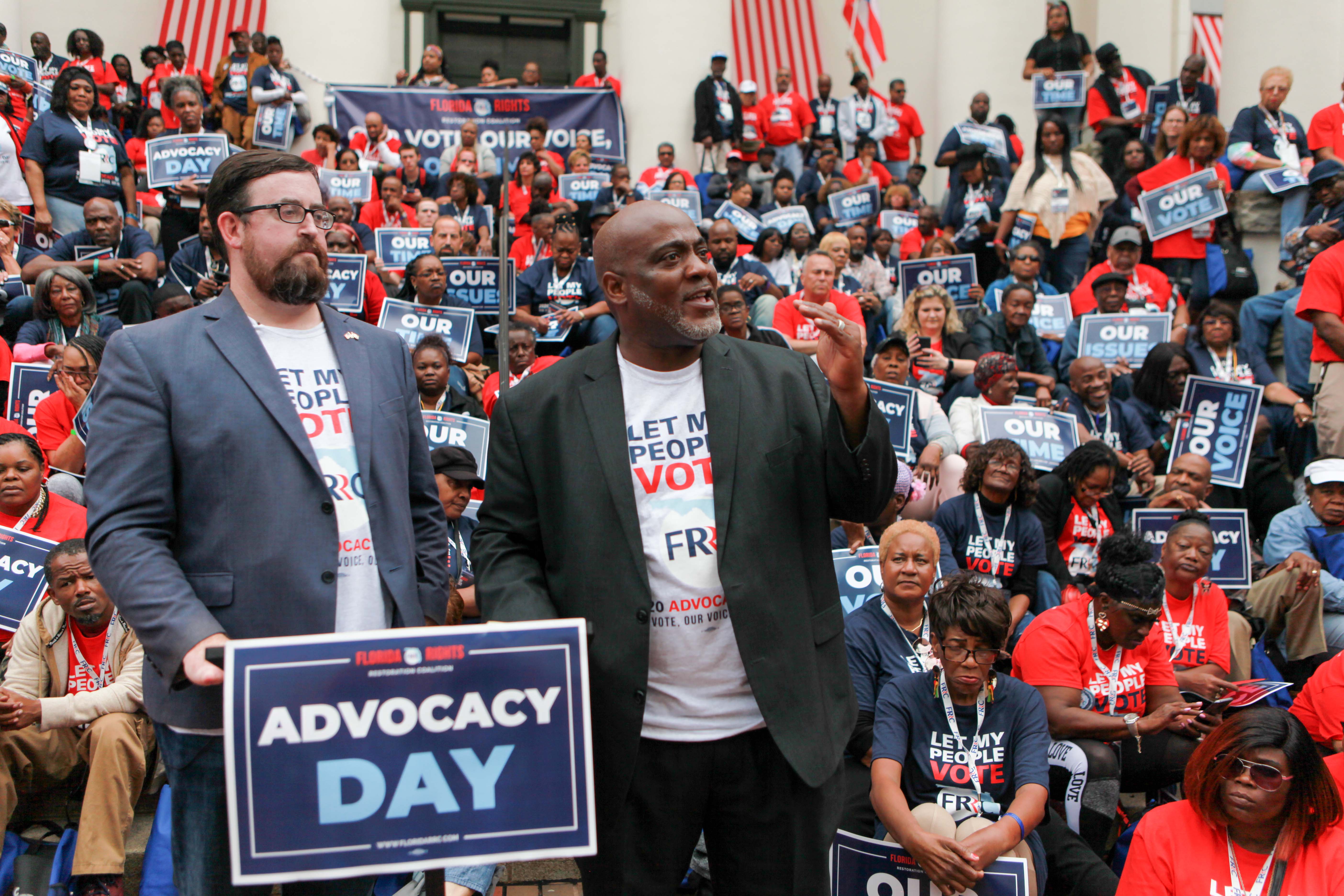 Two speakers at podium in front of large crowd holding signs