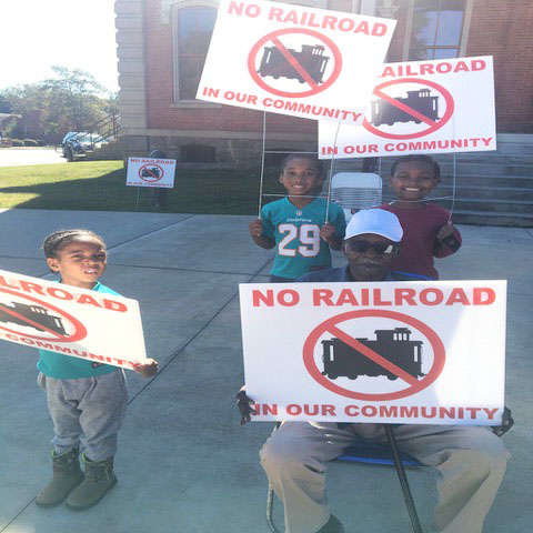 Children and adult hold signs that read "No railroad in our community"