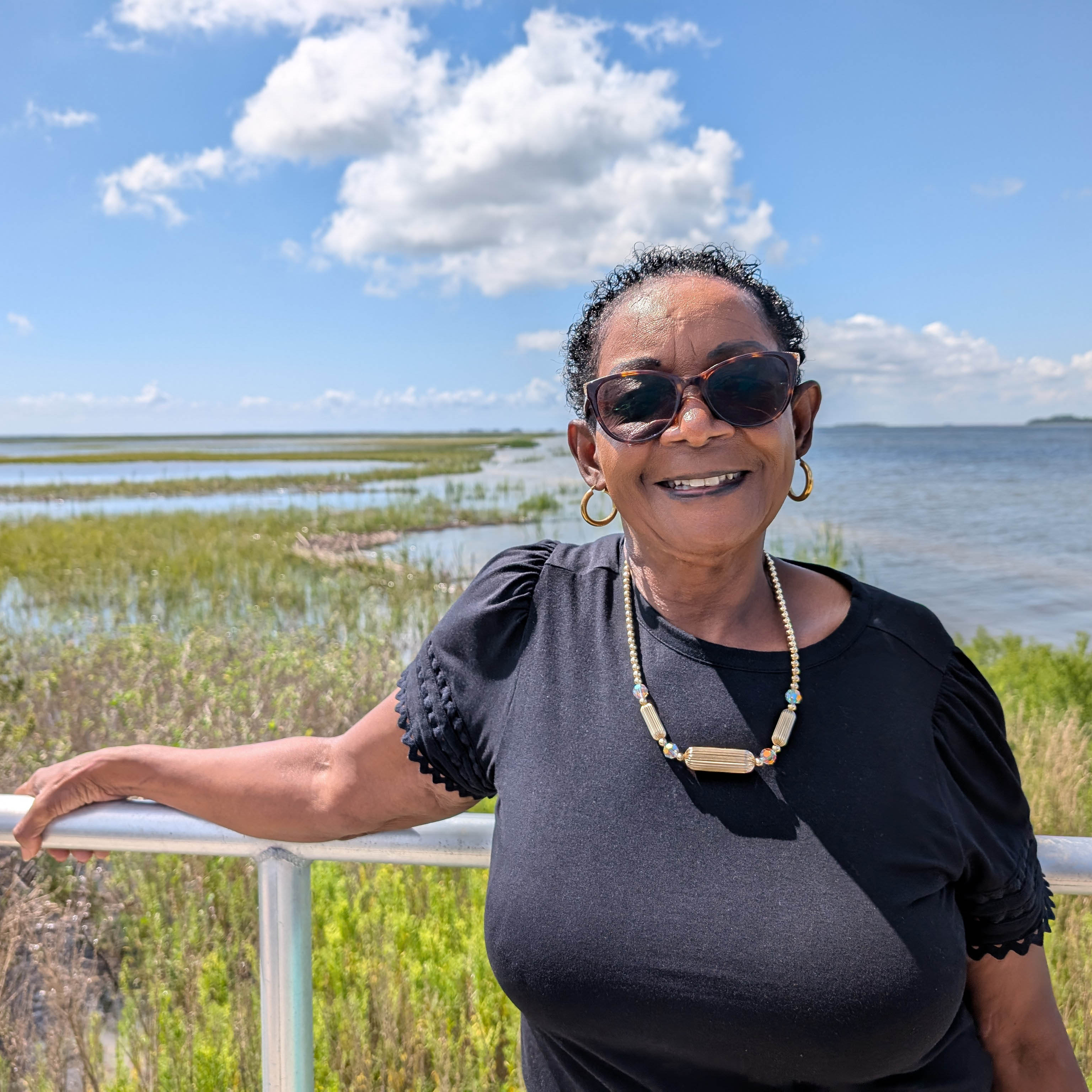Person in sunglasses leans against a railing situated along a shoreline.