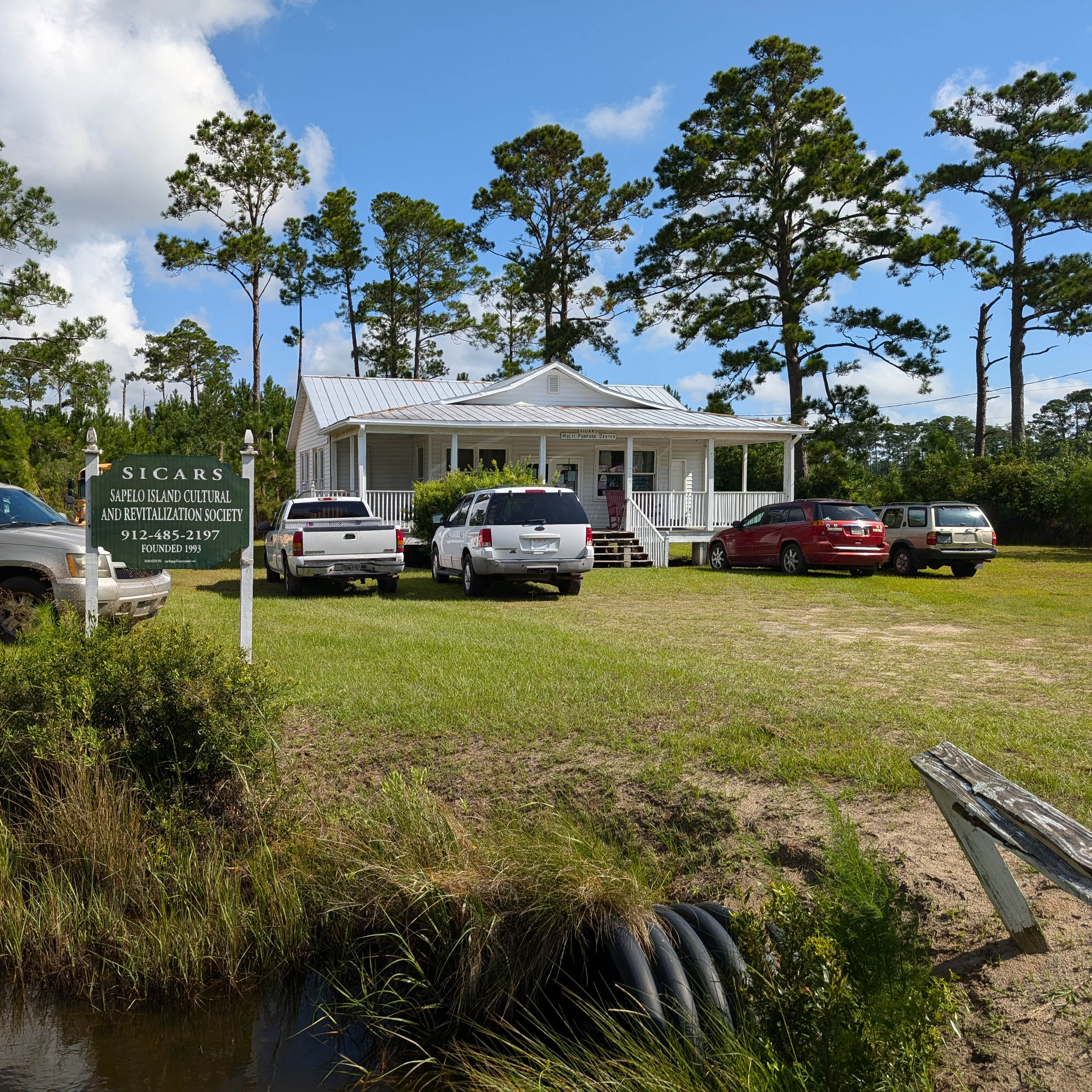 Exterior of building surrounded by large trees on a field of grass occupied by vehicles.