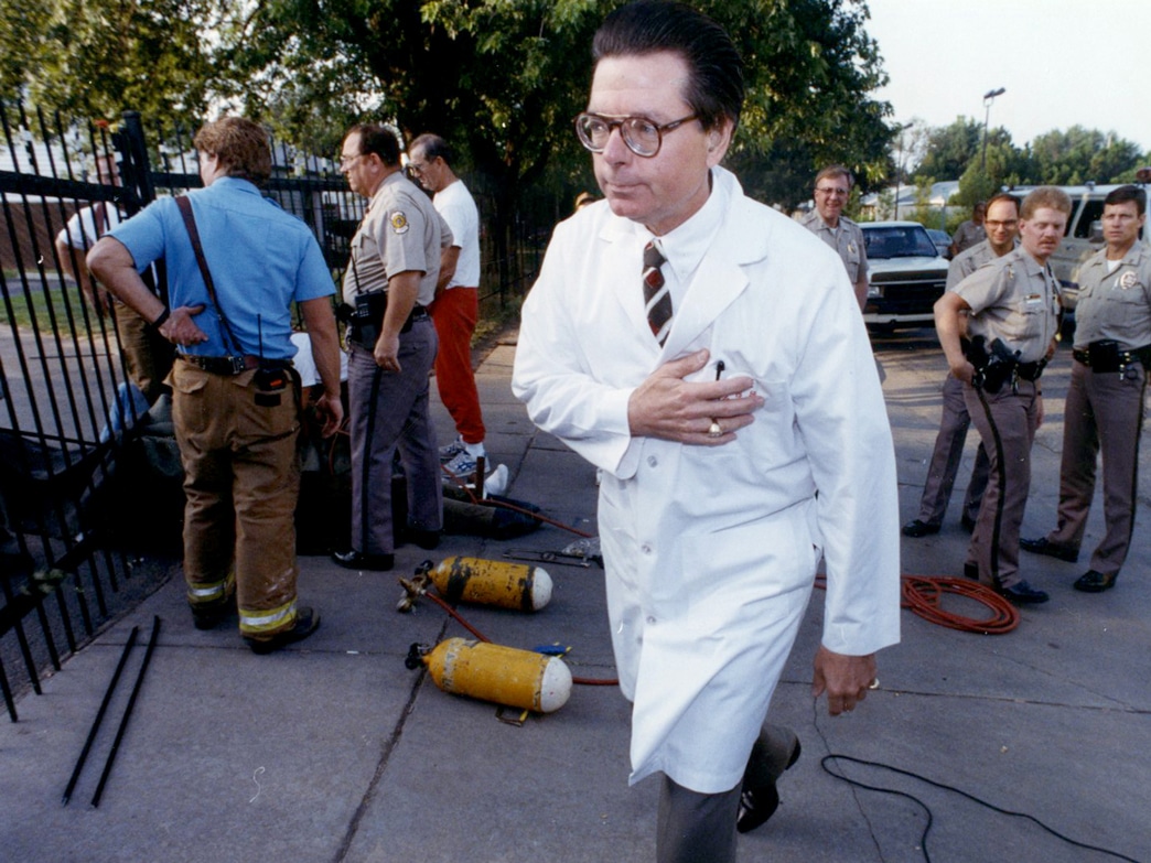 Person in doctor's robes walks past groups of law enforcement officials near a gated area.