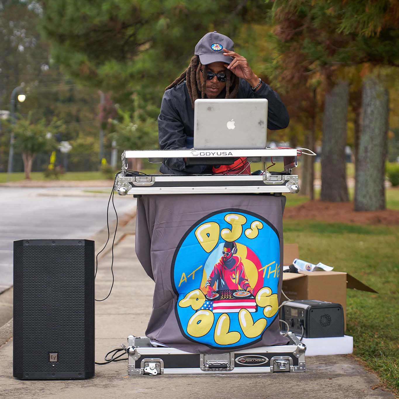 A person in hat and sunglasses behind a stand and speaker looks intently at a laptop programming music.