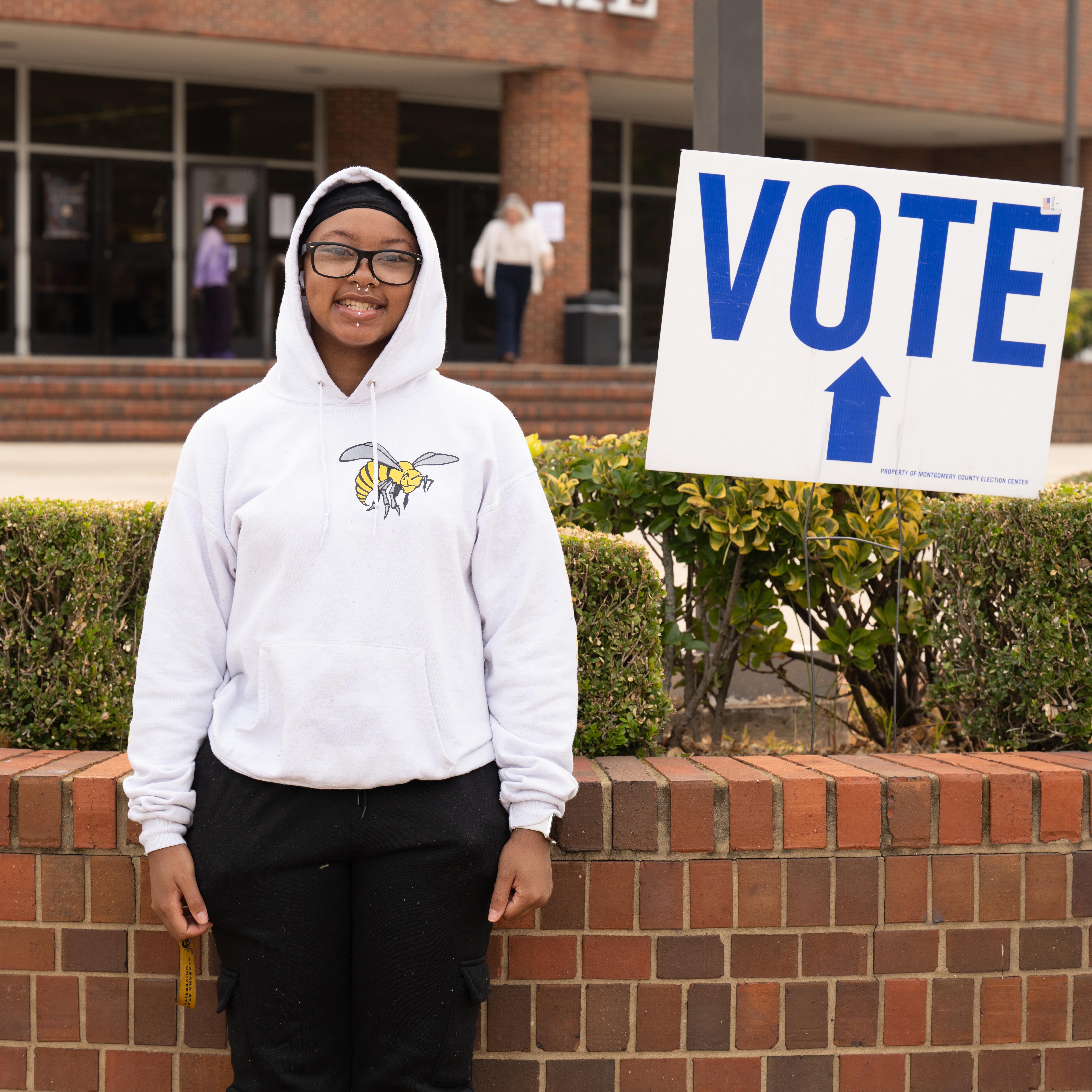 Person in a white hoodie stands outside by a sign pointing to voting place.