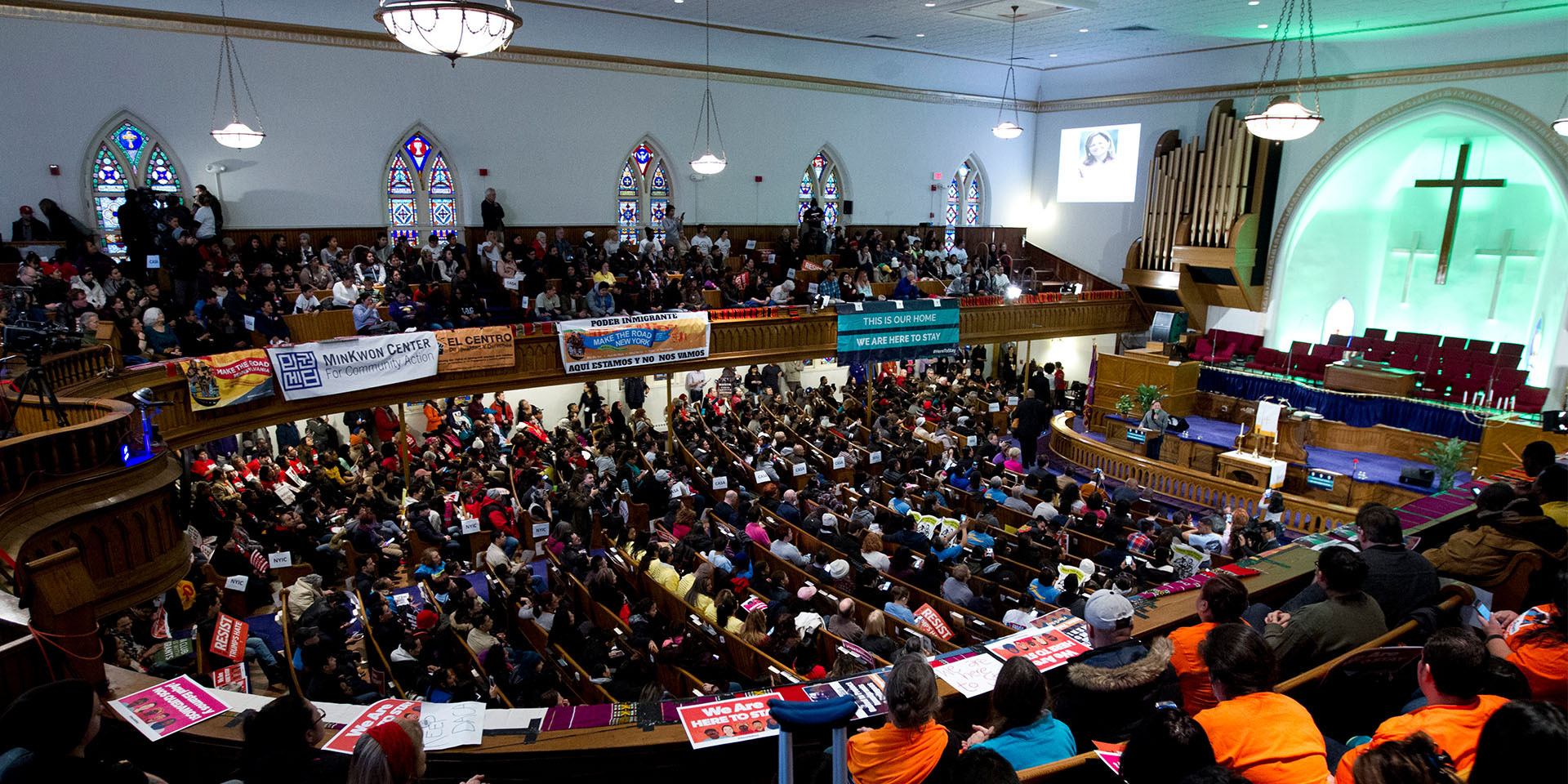 Interior of house of worship in a half circle on two floors with large number of people in pews and banners hanging from the railings.