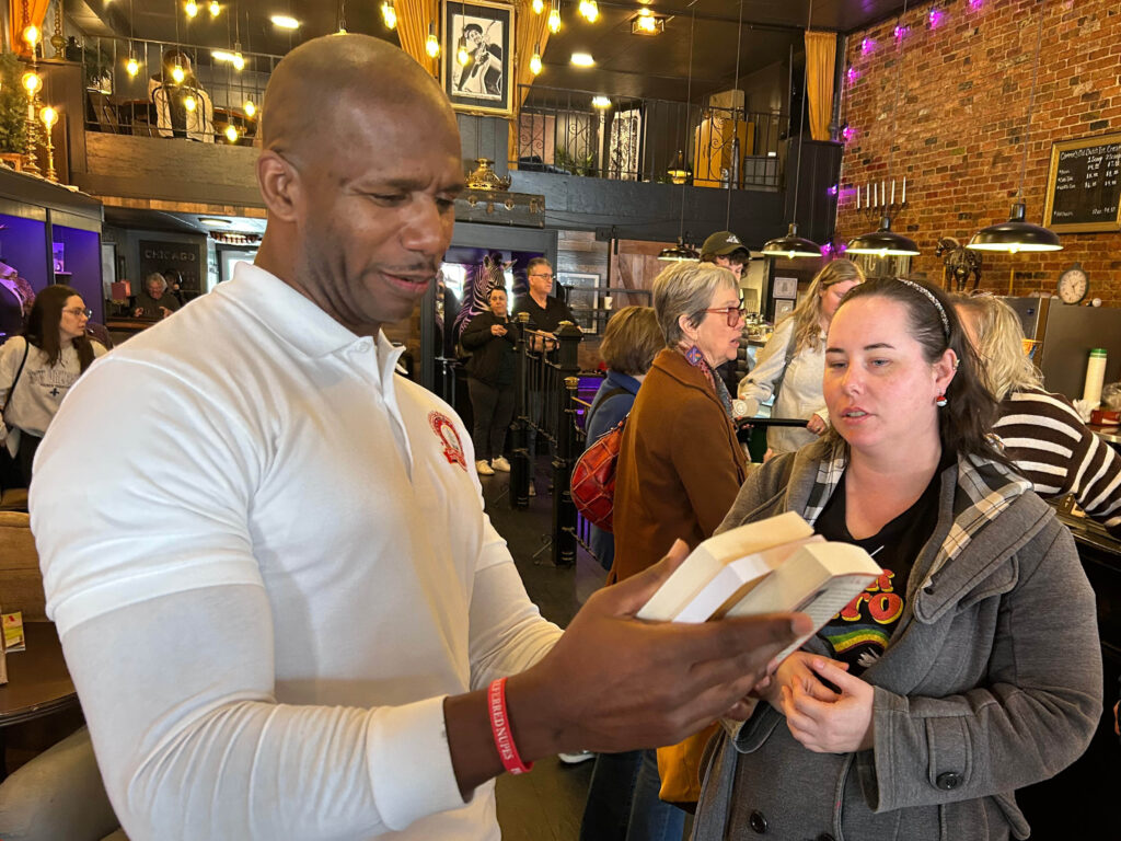 In interior setting with people in chairs a person reviews the back page of a book while engaged in conversation with a person standing nearby.