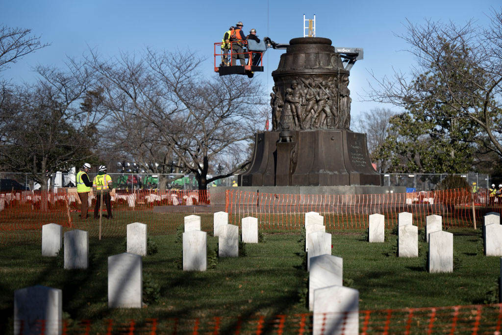 A cherry picker crane with workers rises above a structure one a field with headstones in the foreground.