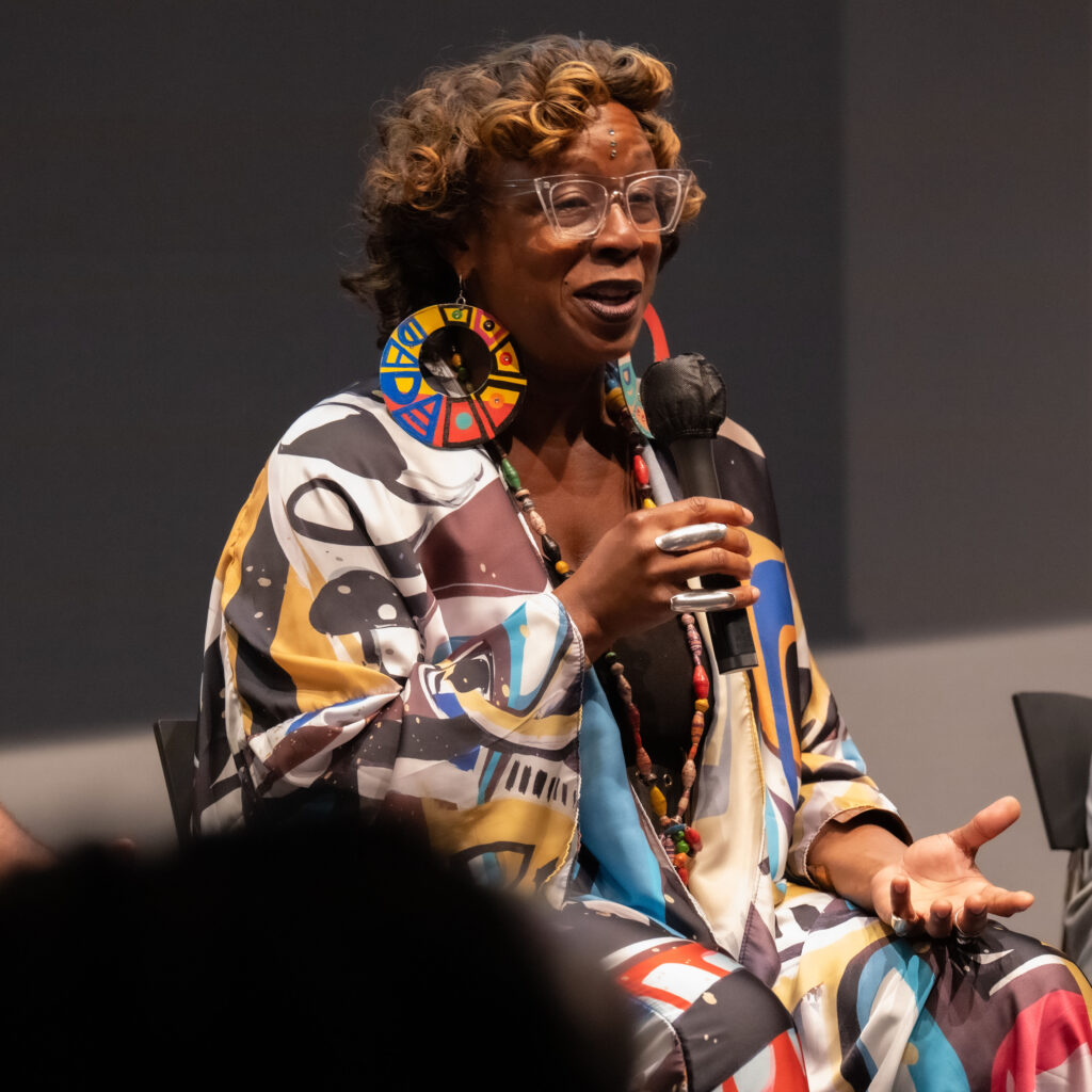 A woman wearing a multi-colored jacket and large circular earrings speaks through a microphone during a panel discussion.