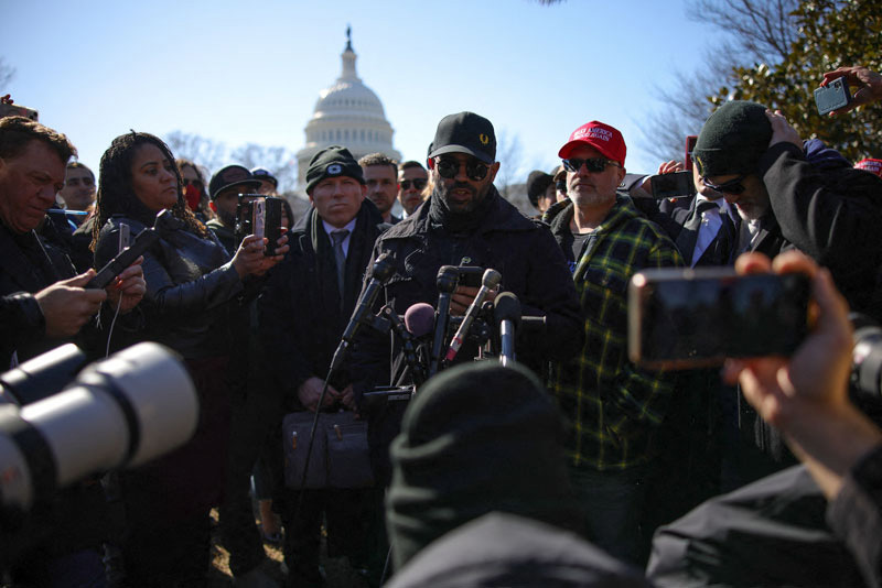 Crowd stands in front of press corp with U.S. Capitol dome far off in background
