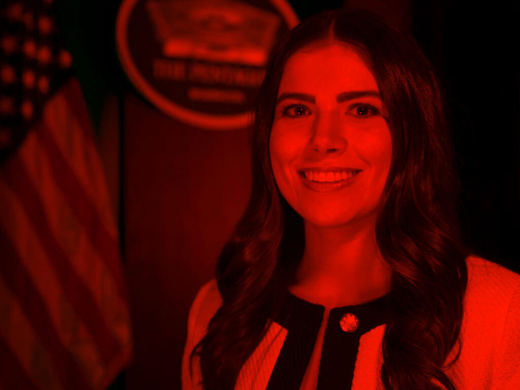 Portrait of person in front of US flag and governmental office seal highlighted by vibrant color.