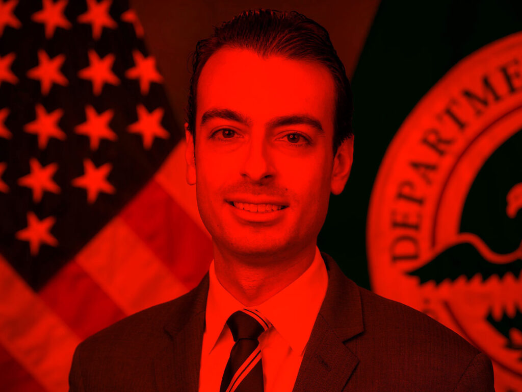 Portrait of person in front of a US flag and government office seal highlighted by vibrant color.