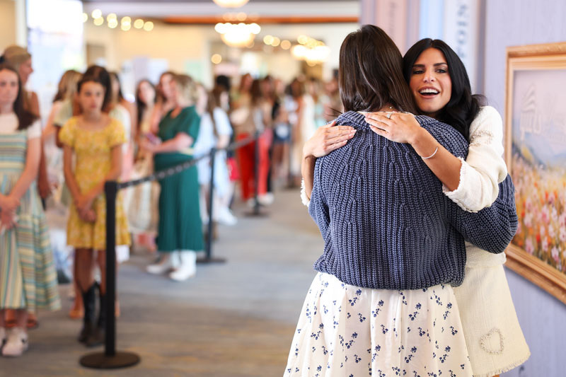 Two people hug - once facing camera - off to the side of a long line taking up a wide hallway.