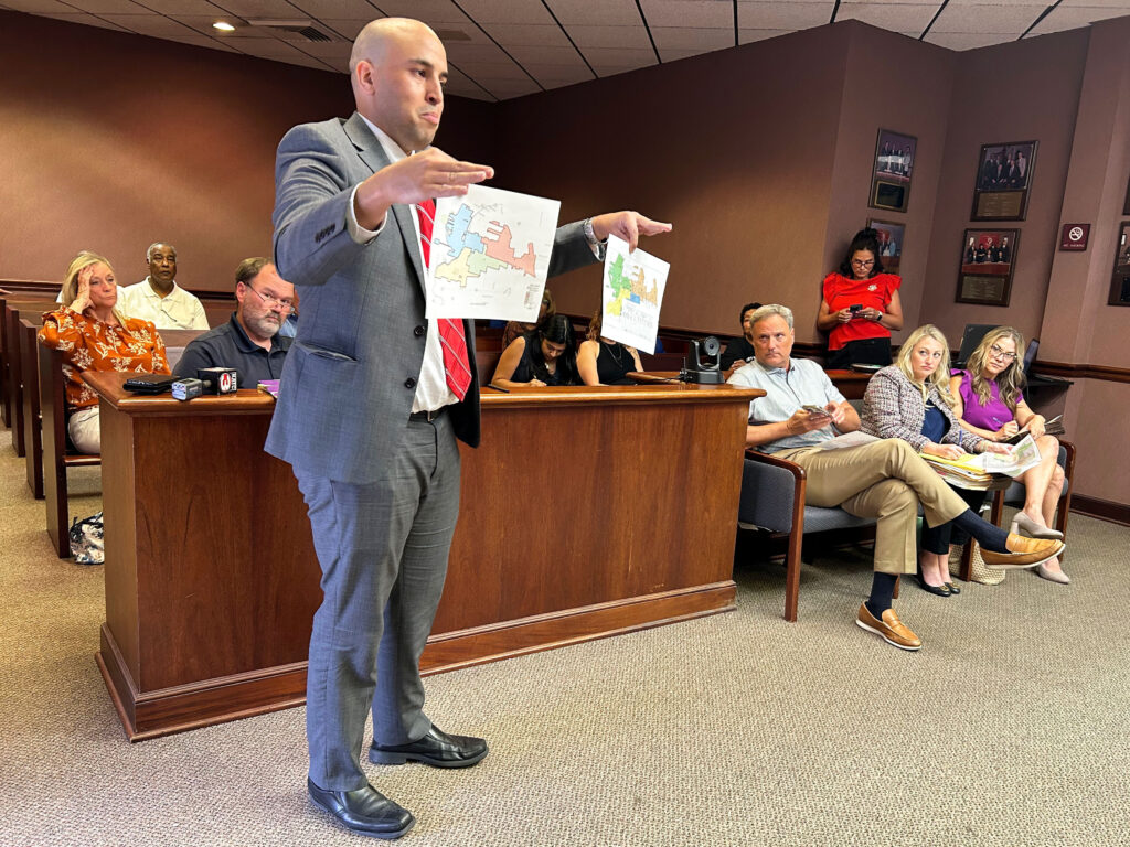 Person stands in front of table holding two pieces of paper showing differing maps.
