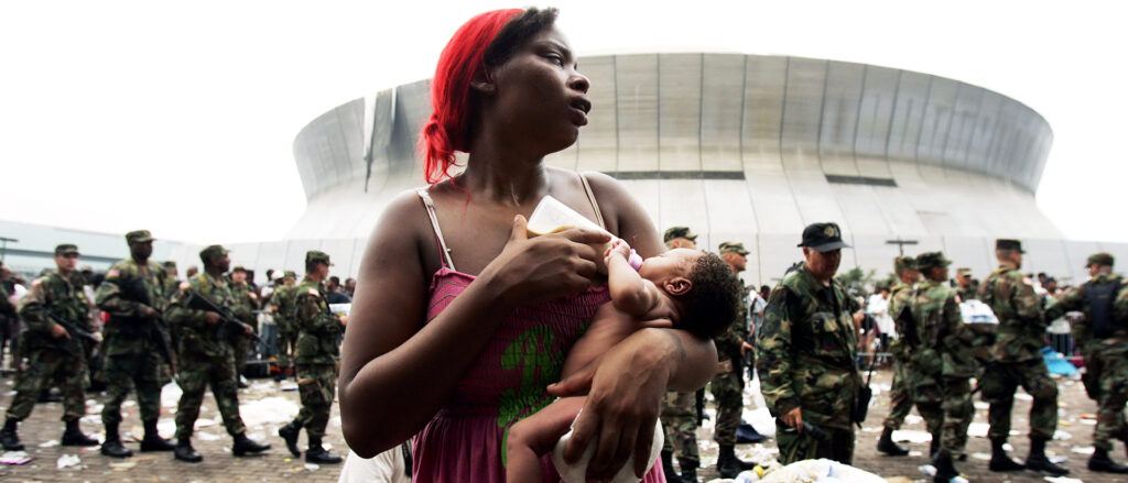 A person feeds a child through a bottle as personnel in military attire line up in the background outside a stadium facility.