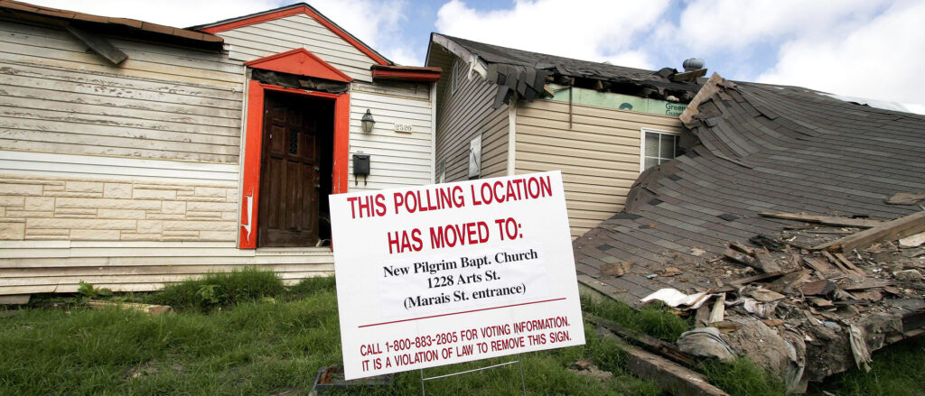 A sign alerting of polling station change sits in front of a collapsed building.