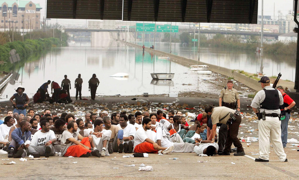 Large group of people seated on a roadway away from flooded waters.
