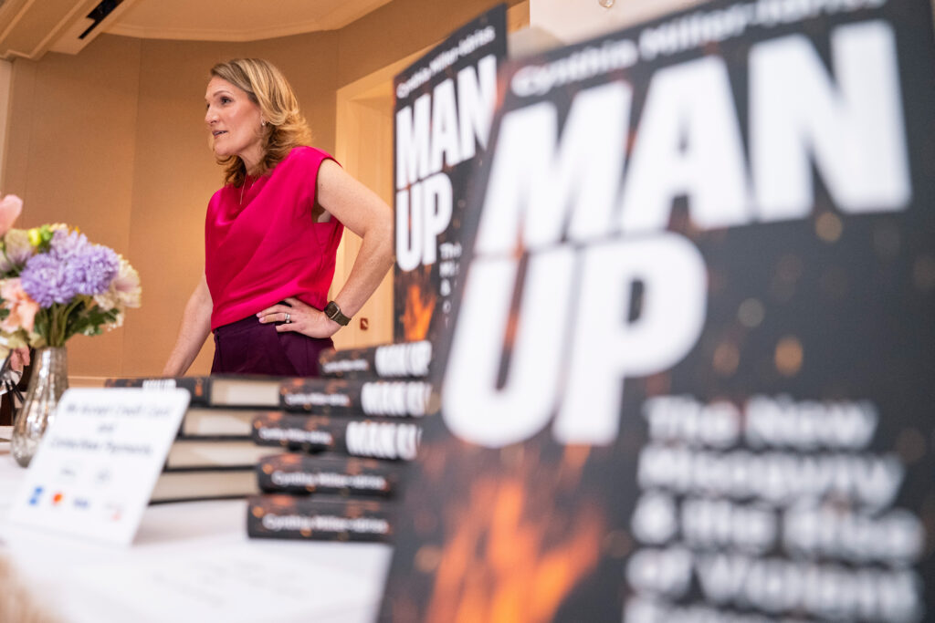 person in a red shirt next two a table stocked with books entitled "Man Up"