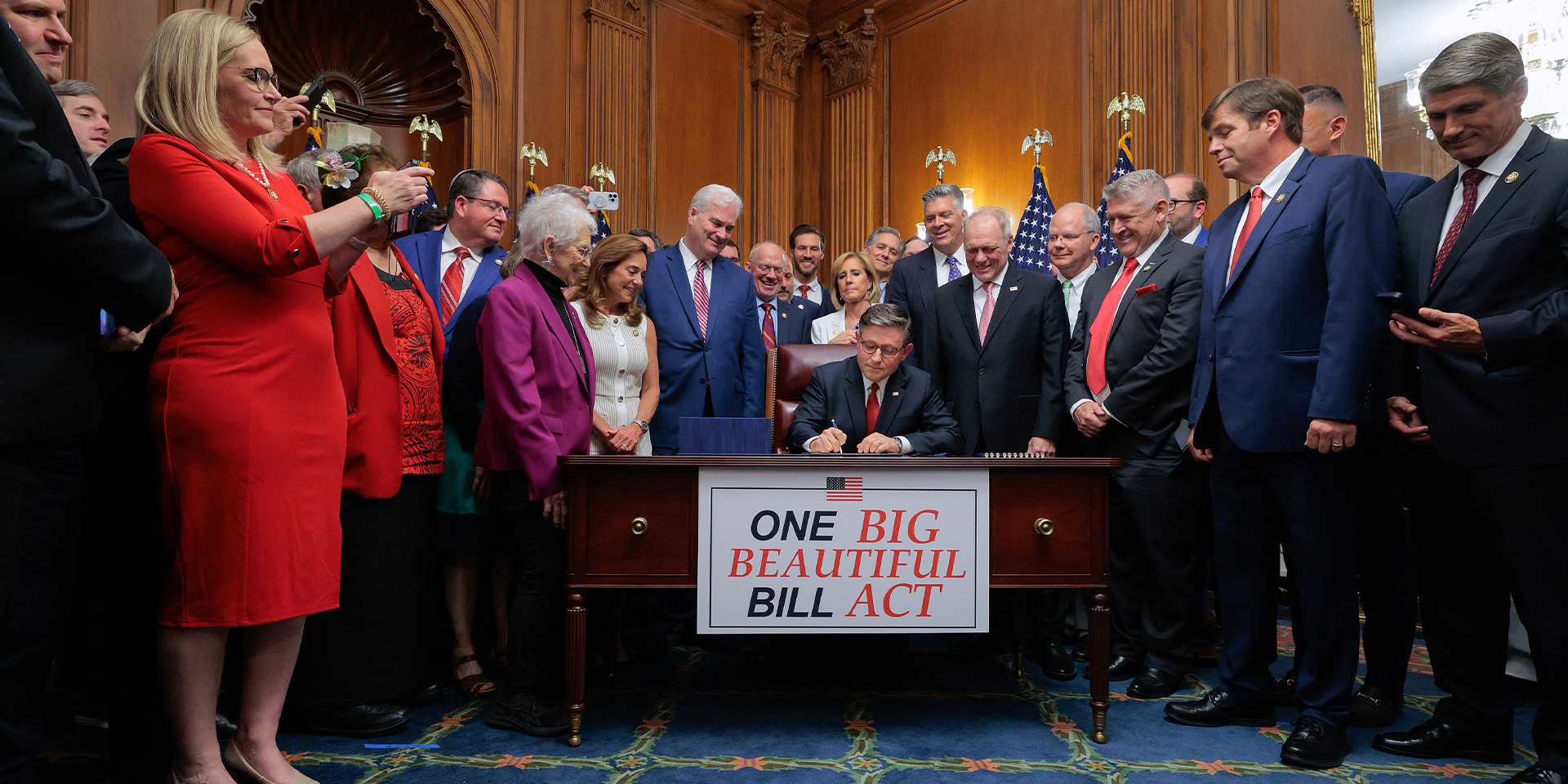 Person at desk using pen to sign document is surrounded by other people, one taking a photo with a phone.
