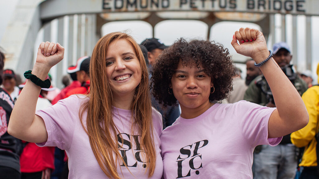 Two people on a crowded bridge raise their their fists. They are wearing pink shirts that say, 'SPLC.'
