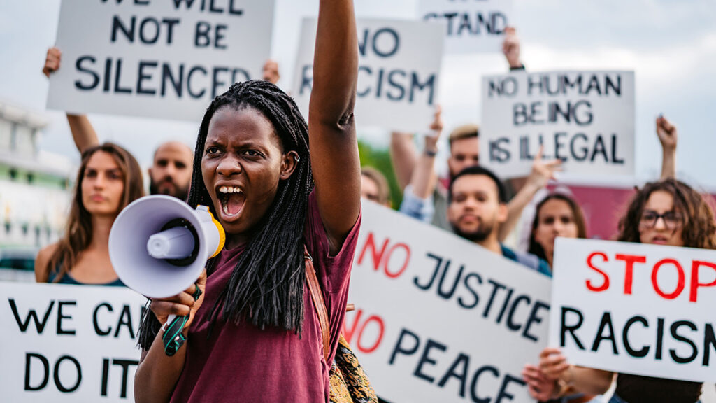 A protester holds a megaphone and raises a fist. Others in the background hold numerous signs.