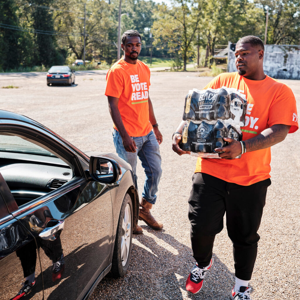 Two volunteers in orange shirts that say 'Be Vote Ready.' One is carrying two packages of water bottles.