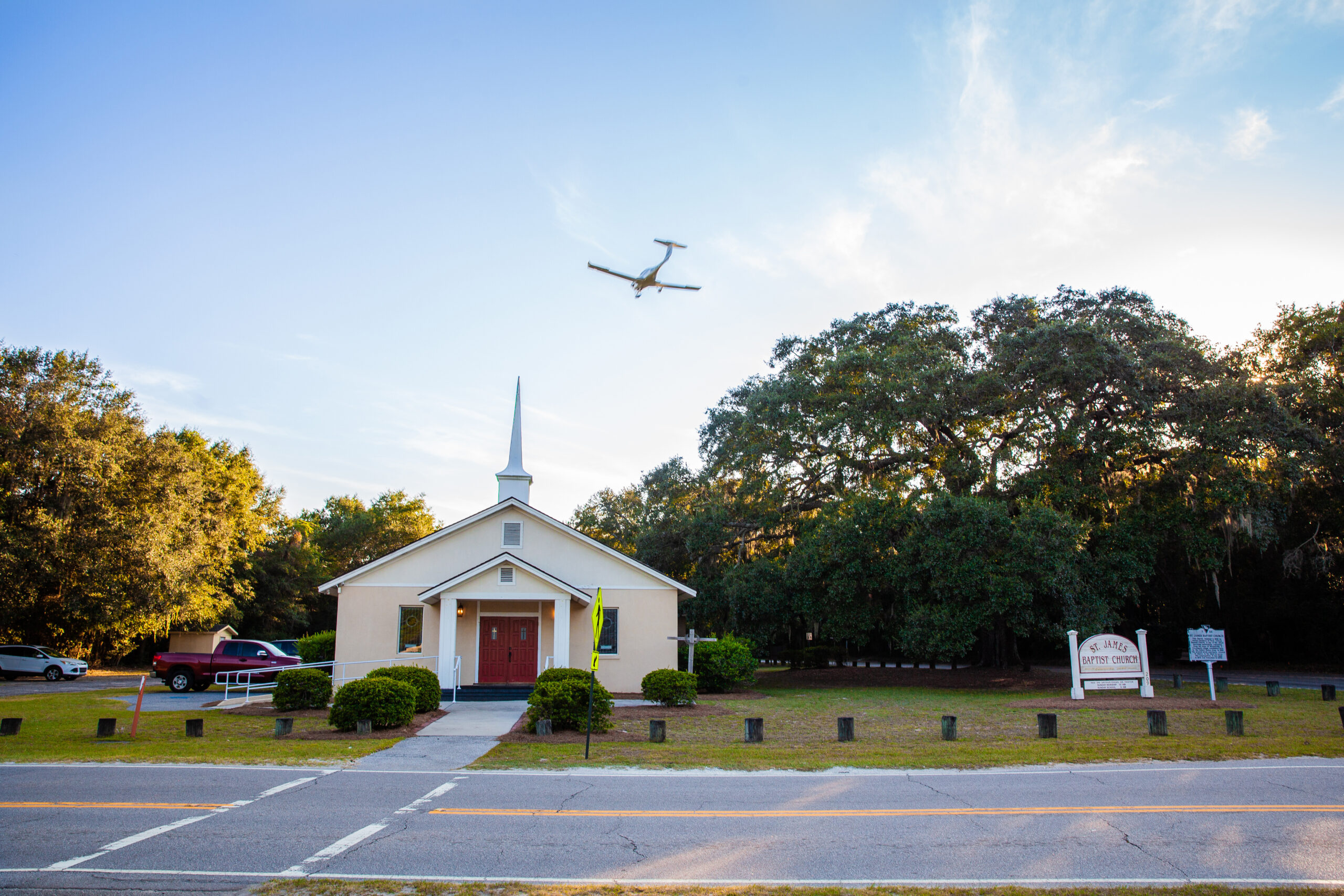 A white church with a red door in front of an empty parking lot. A plane flies overhead.
