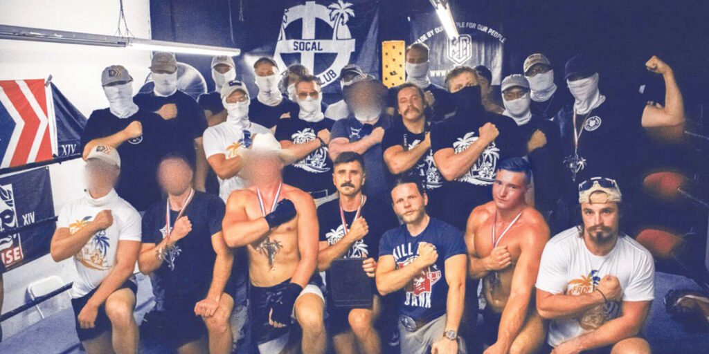 Large group of people posing standing in kneeling with right hand formed in a fist over chest in an interior space with banners in background.