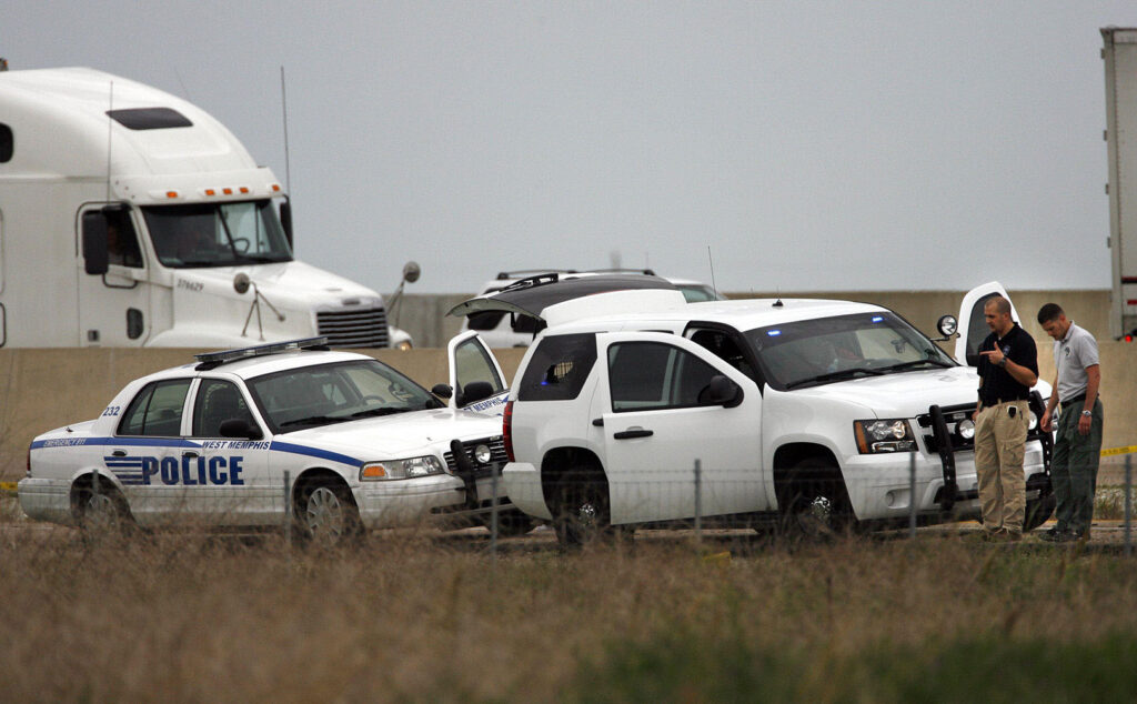 People hold a conversation in front of two cars on the side of a road with heavy traffic.