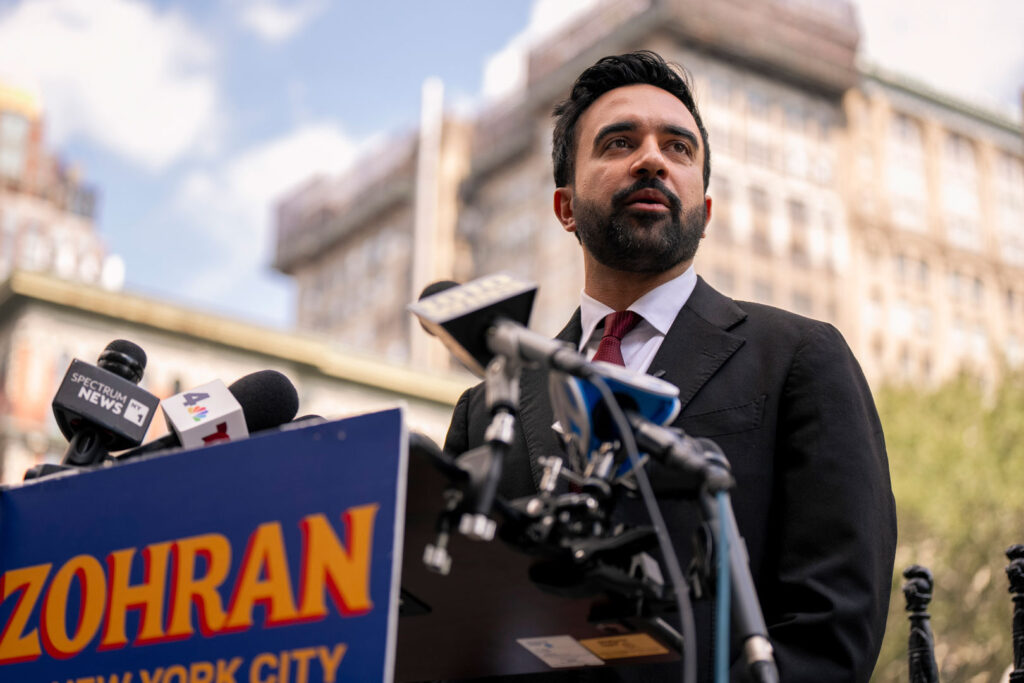 Person speaking at podium with numerous microphones and political signage on front in an exterior setting.