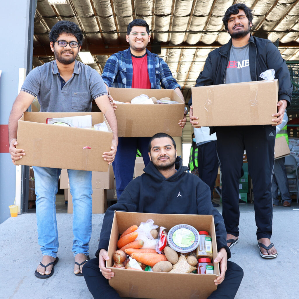 Four people sitting and standing hold boxes of goods in exterior setting.