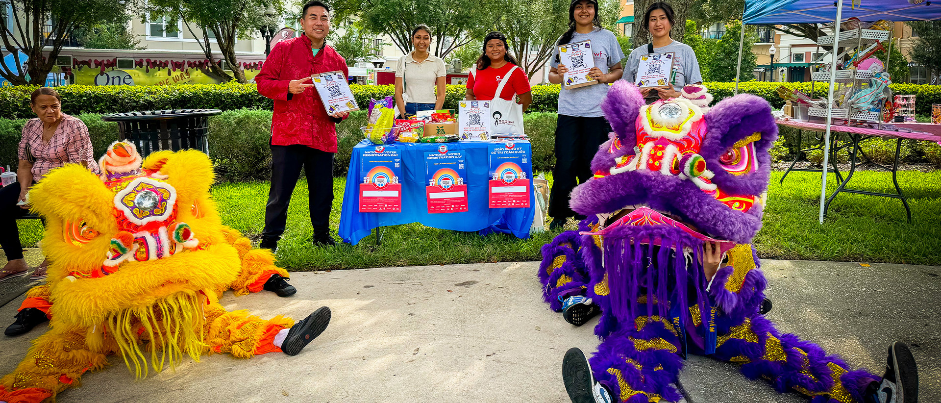 Costumed dragons in vibrant colors before a group near a table in exterior setting.