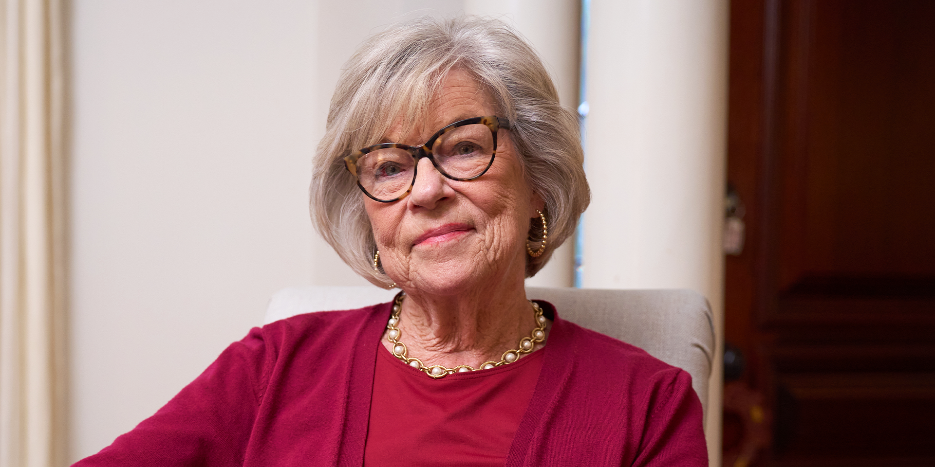 An elderly woman in a red shirt with a pearl necklace.