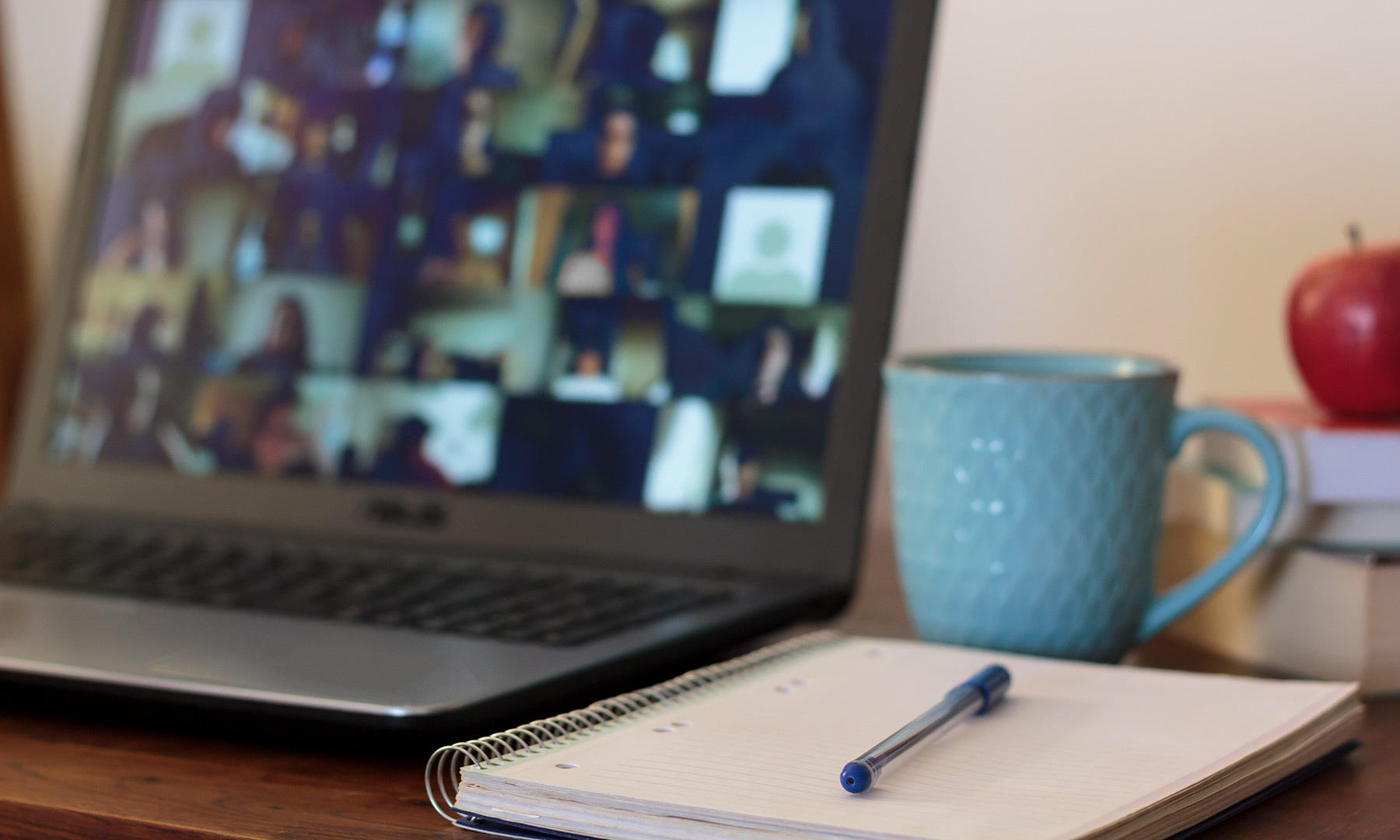 Computer, notebook, pen, coffee cup sit on a desk with a stack of books and an apple on top.