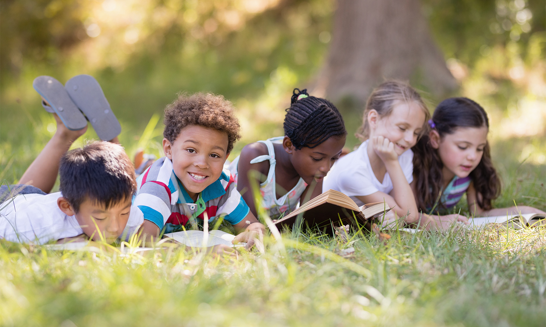 A group of children lie on their stomachs in the grass reading books in front of a tree on a sunny day. One child looks directly at the camera and smiles.