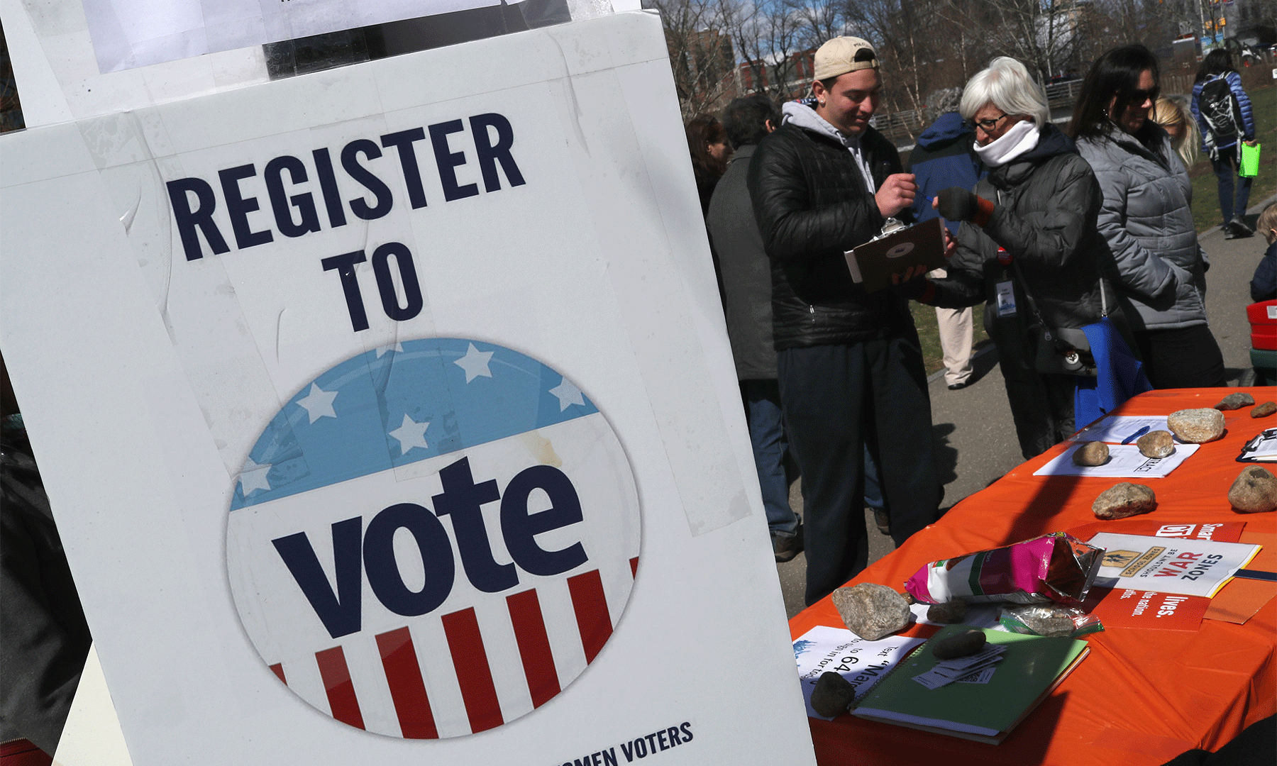 Piece of paper taped to a column that says "Register to Vote" with several people standing in the background interacting with each other.