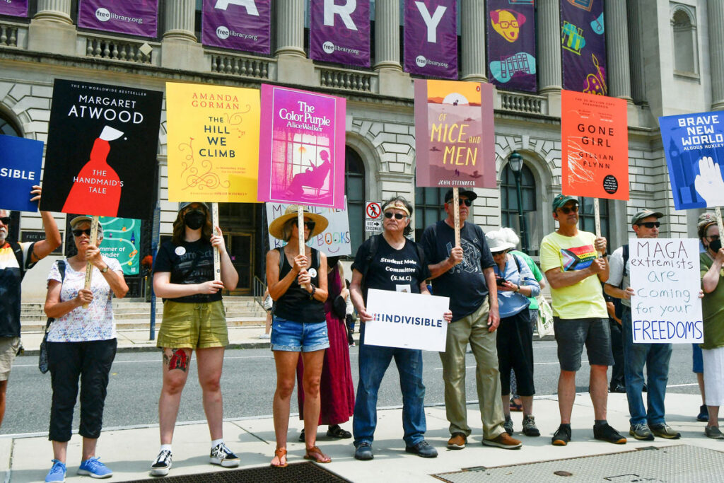 Group of people lined up outside a library building holding signs featuring the cover of banned books.