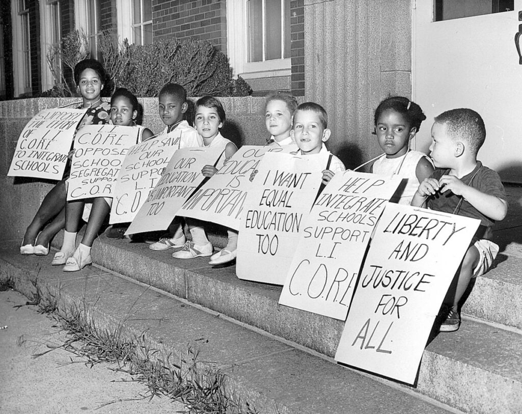 Children hold large signs in front of them as they sit on short staircase in an outdoor setting.