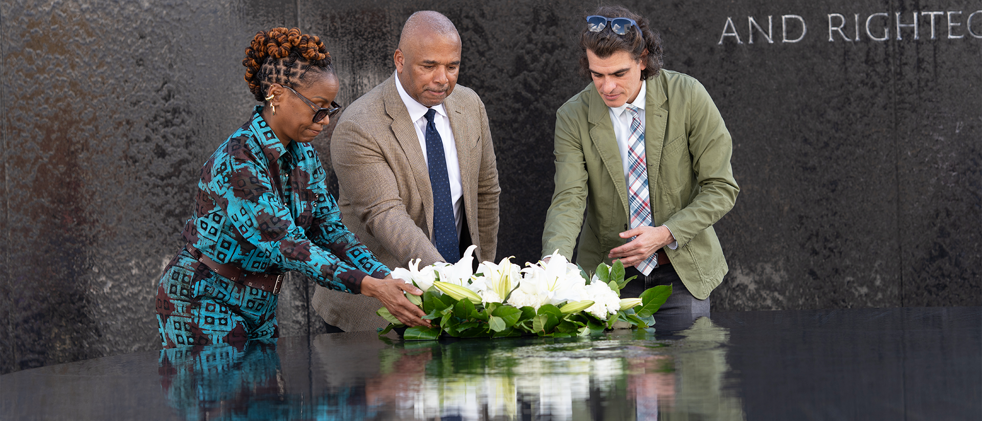 Three people hold a flower arrangement over a water sculpture in an outdoor setting.